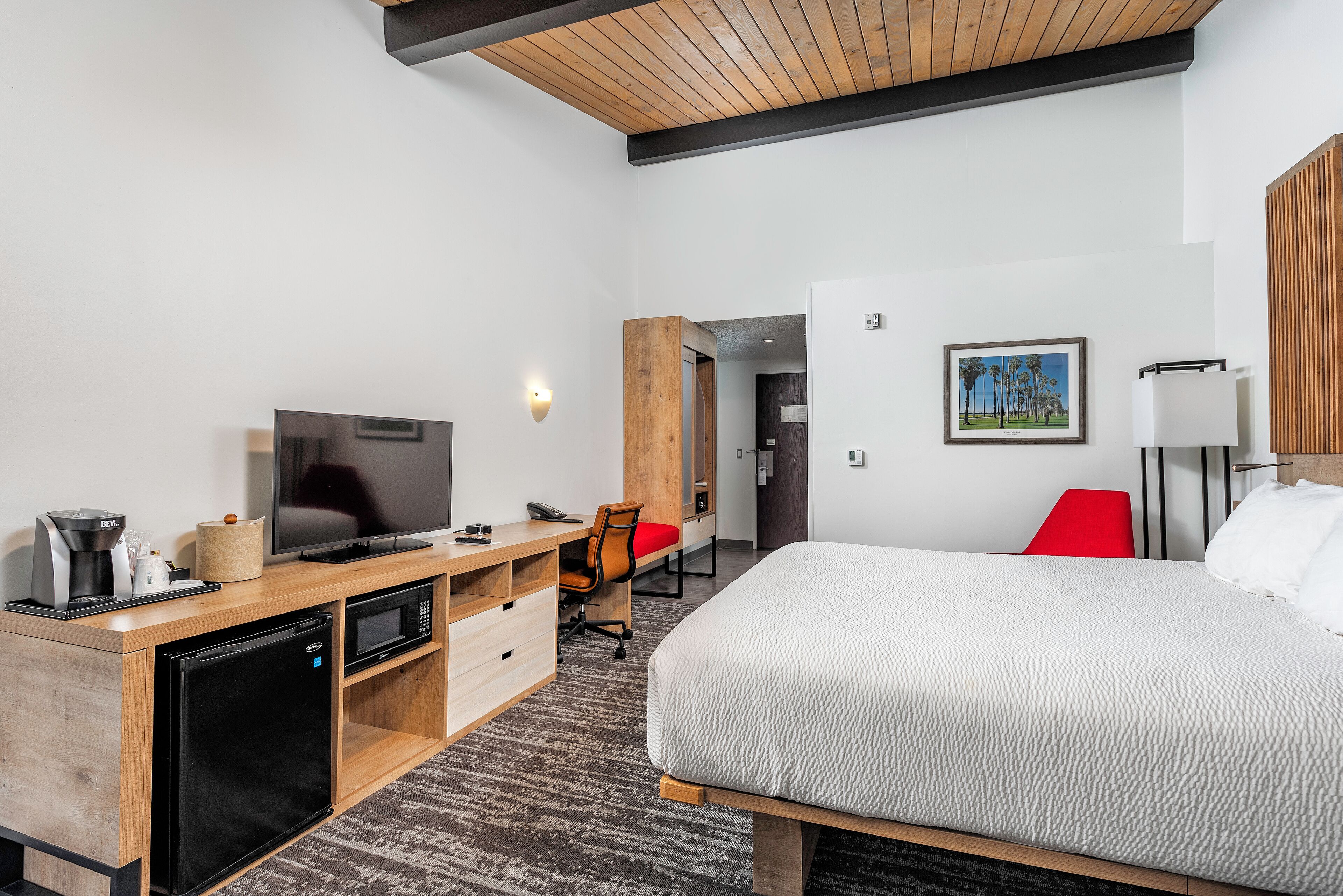 Habitación de hotel con cama, escritorio con televisor, refrigerador, microondas, silla y un cuadro en la pared.