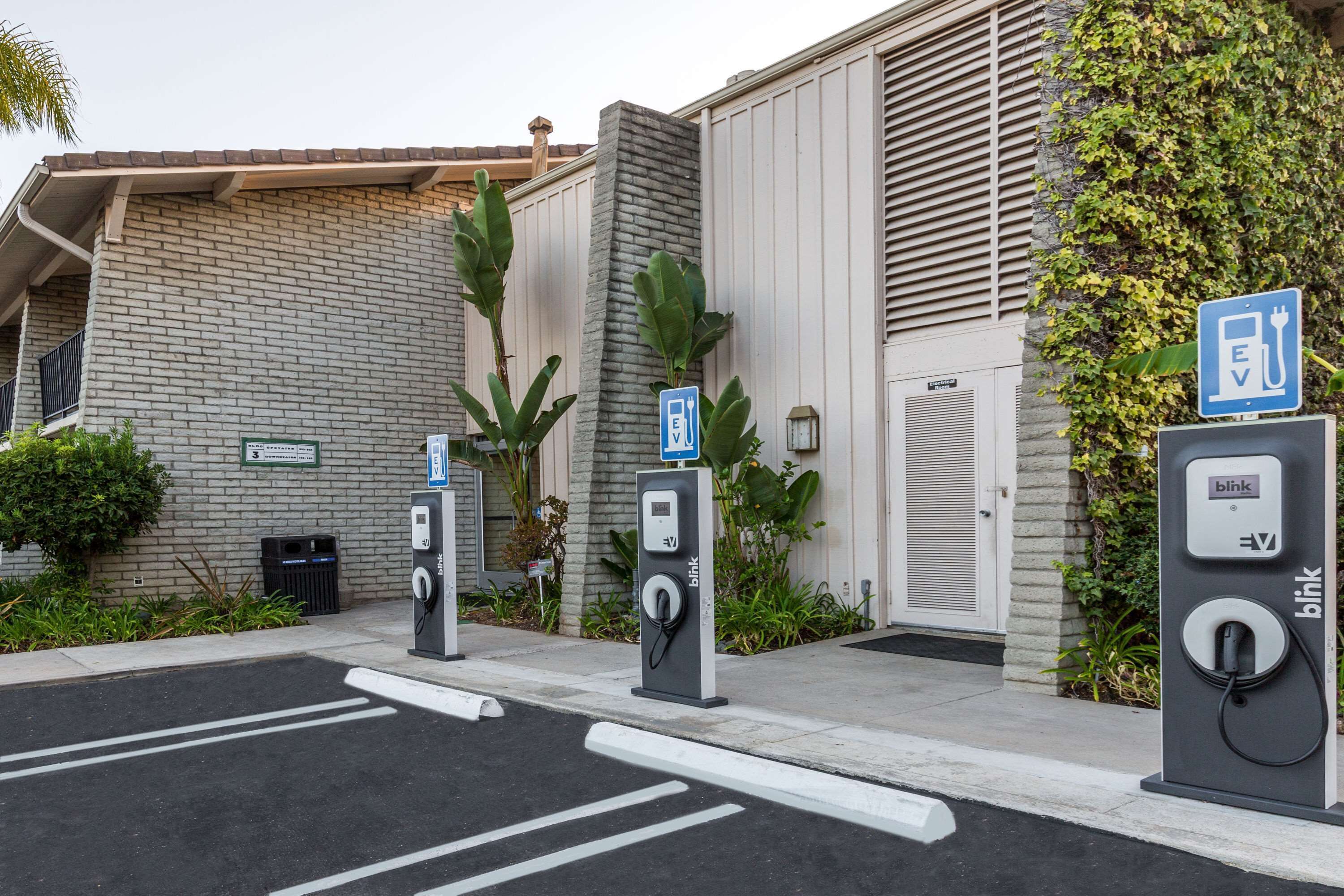 An electric vehicle charging station area with three charging units, a brick building, and a white door.