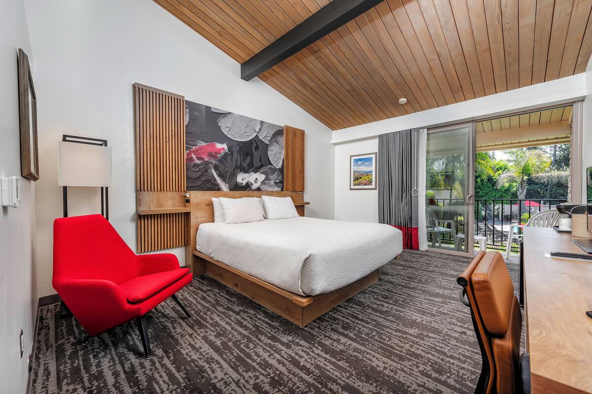A modern hotel room with a large bed, a red armchair, a wooden headboard, and a large window offering a view of the outdoors.