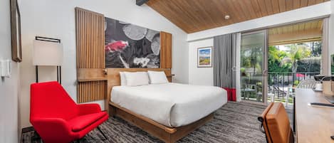 A modern hotel room with a large bed, a red armchair, a wooden headboard, and a large window offering a view of the outdoors.