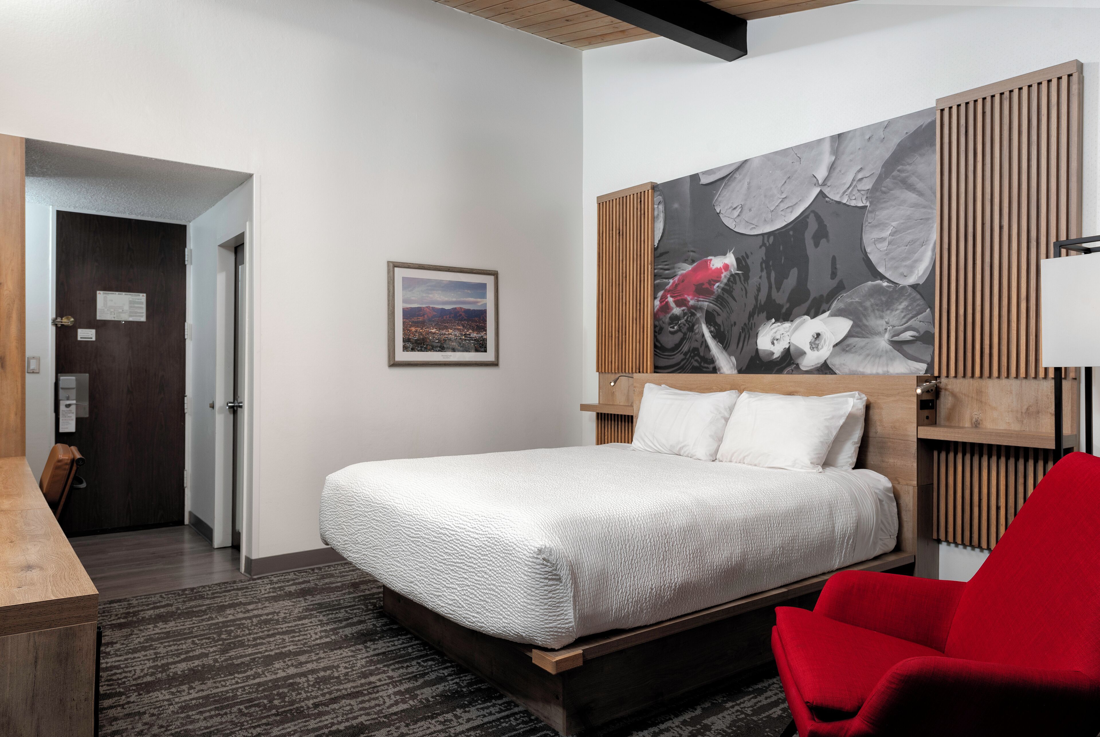 Quarto de hotel com cama, uma cadeira vermelha, cabeceira de madeira, um quadro na parede e vista da cidade pela janela.