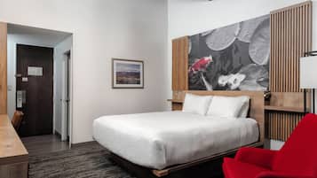 Quarto de hotel com cama, uma cadeira vermelha, cabeceira de madeira, um quadro na parede e vista da cidade pela janela.