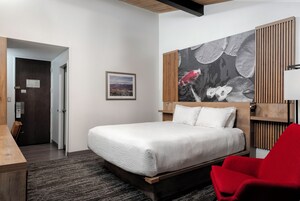 Quarto de hotel com cama, uma cadeira vermelha, cabeceira de madeira, um quadro na parede e vista da cidade pela janela.