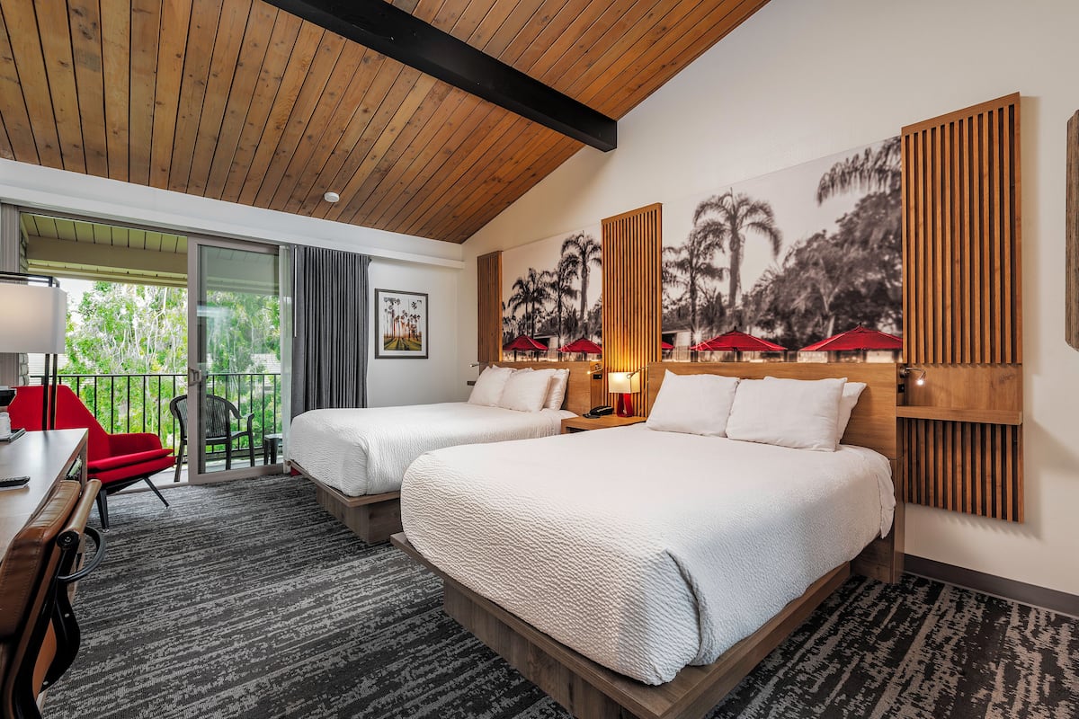 A hotel room with two beds, a desk, a chair, and a balcony with red umbrellas.