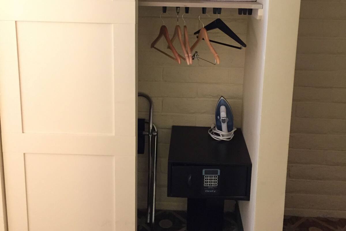 A hotel room with a wardrobe, a small shelf with towels, a hanging rack with hangers, an iron, and a suitcase.