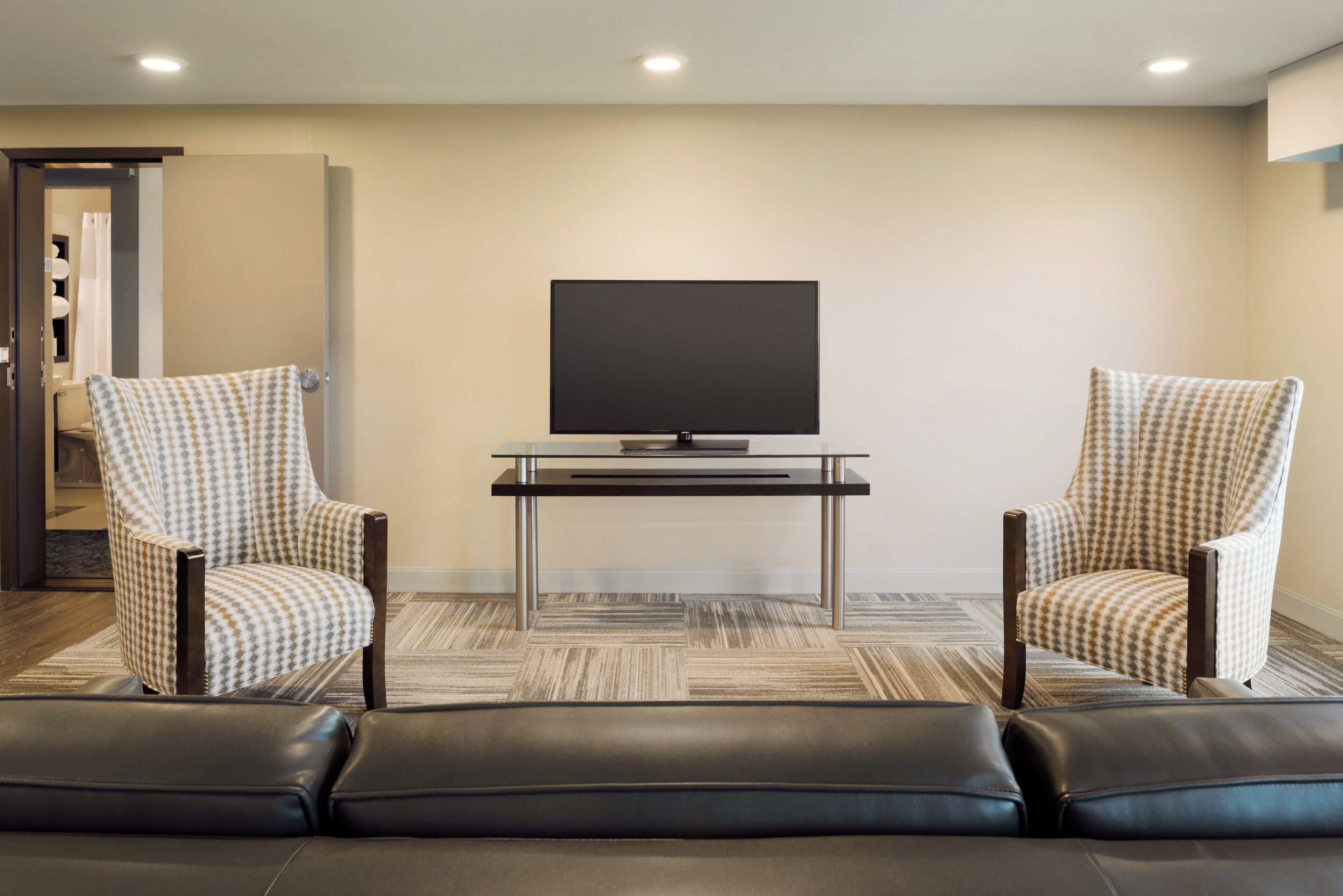 A modern living room with a flat-screen TV, two patterned armchairs, a glass coffee table, and a leather sofa.