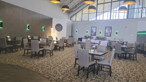 A spacious indoor area with multiple tables and chairs, a patterned carpet, and a high ceiling with natural light. - Holiday Inn Bloomington - Normal by IHG (Normal)