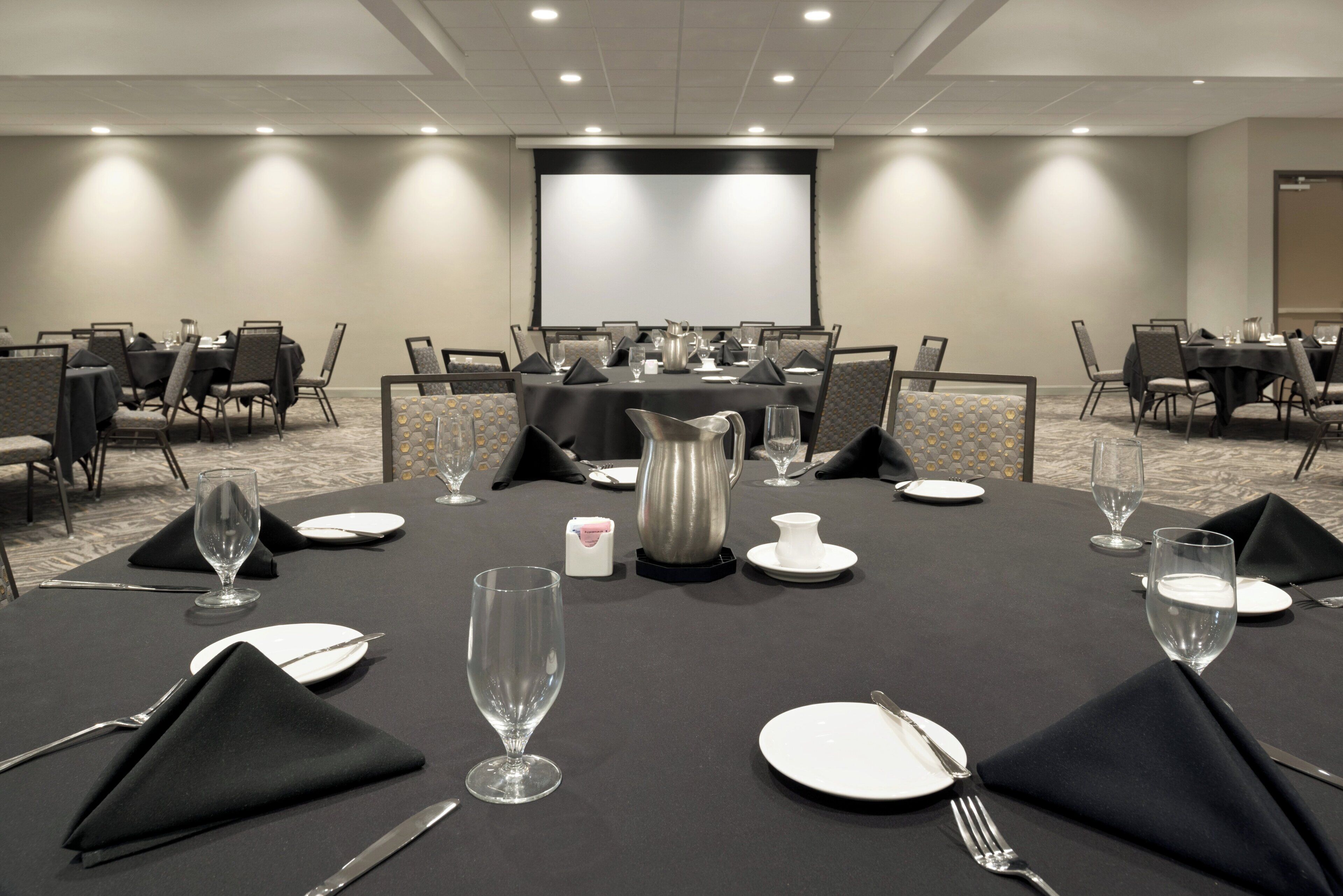 Un salón de conferencias con mesas dispuestas para una reunión, equipado con proyectores y una pantalla.