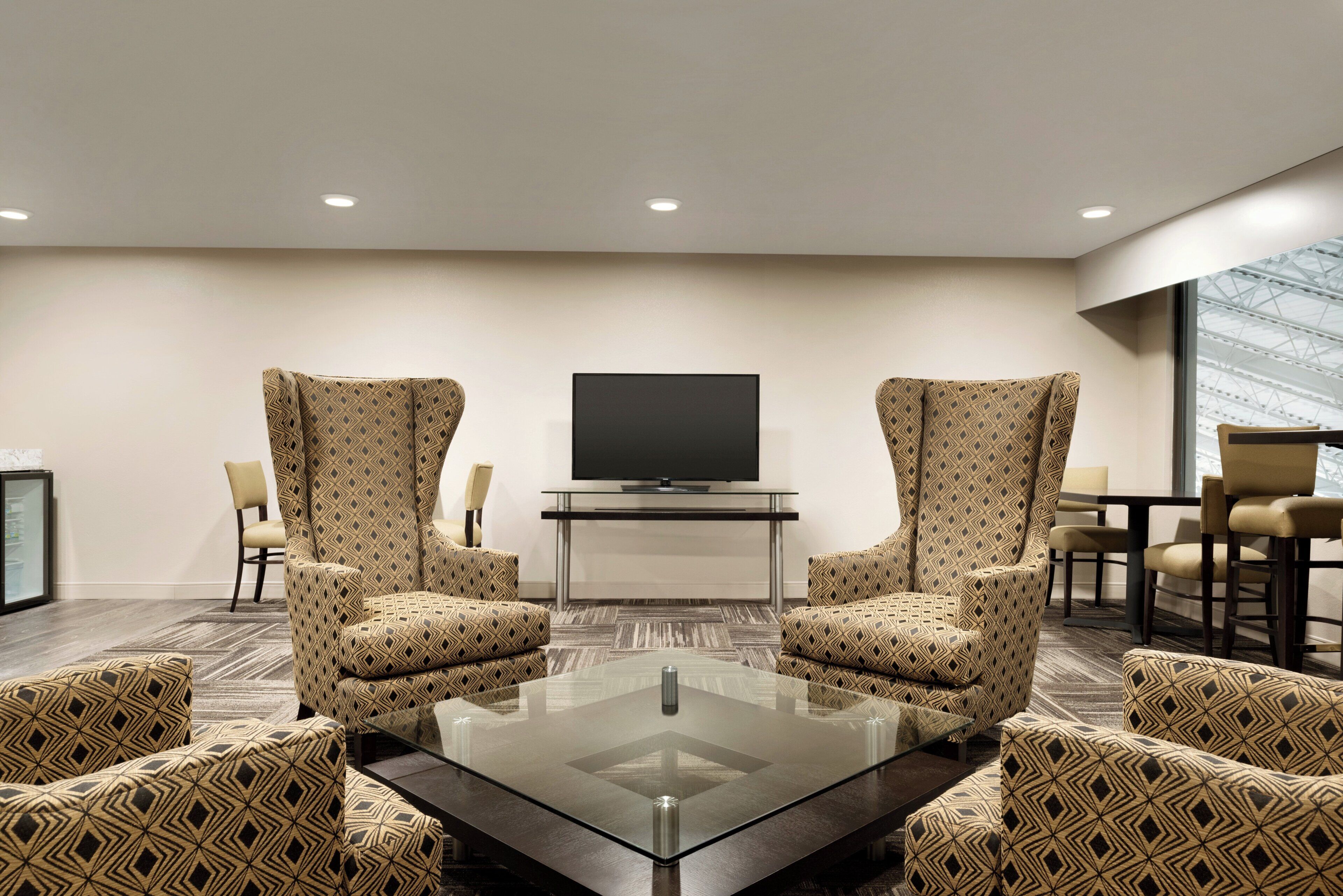 A hotel room with a glass coffee table, two patterned armchairs, a flat-screen TV on a stand, and a view of the sky through a large window.