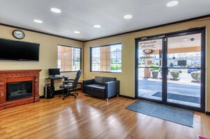 A hotel lobby with a fireplace, a television, a sofa, a desk with a computer, and large windows offering a view of the parking lot.