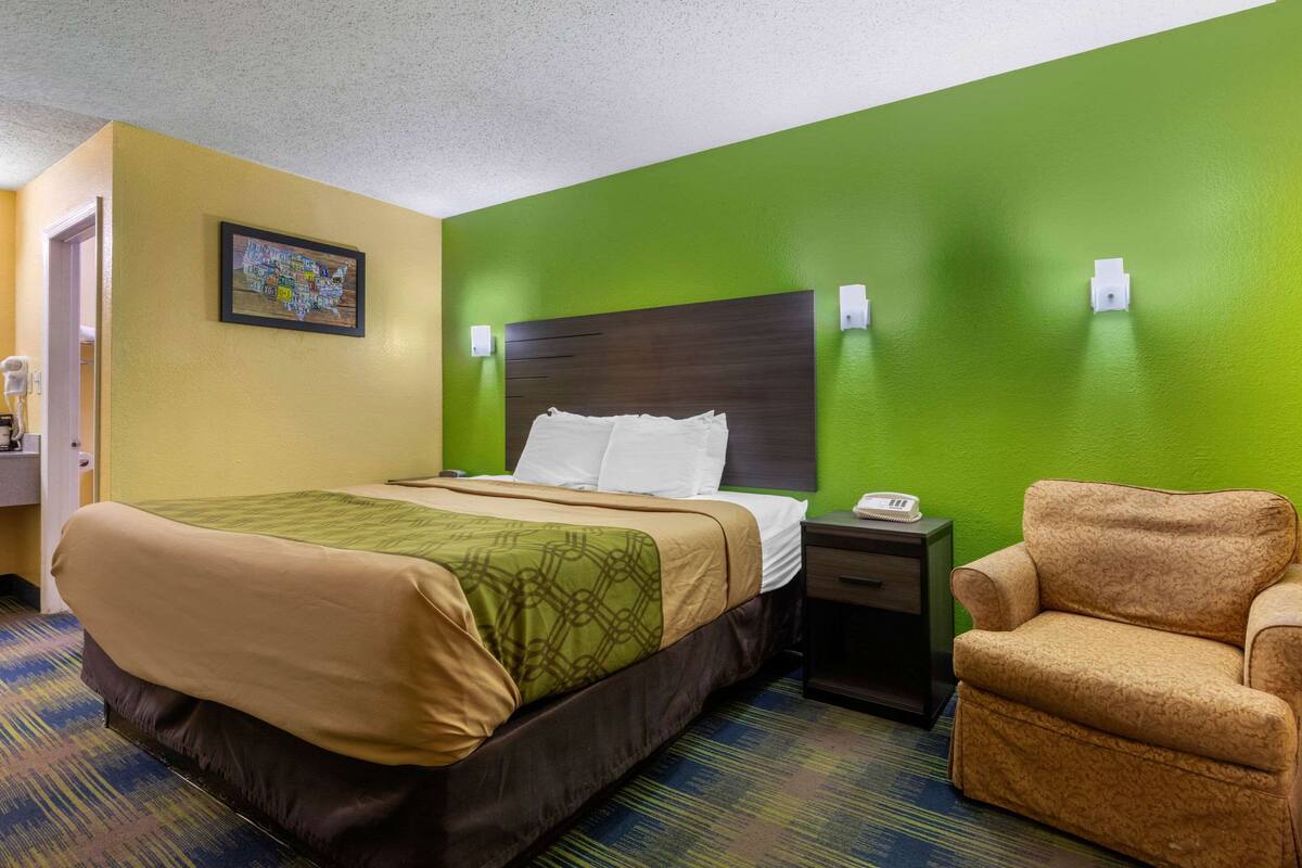 A hotel room with a bed, a chair, a nightstand, and a wall-mounted phone.