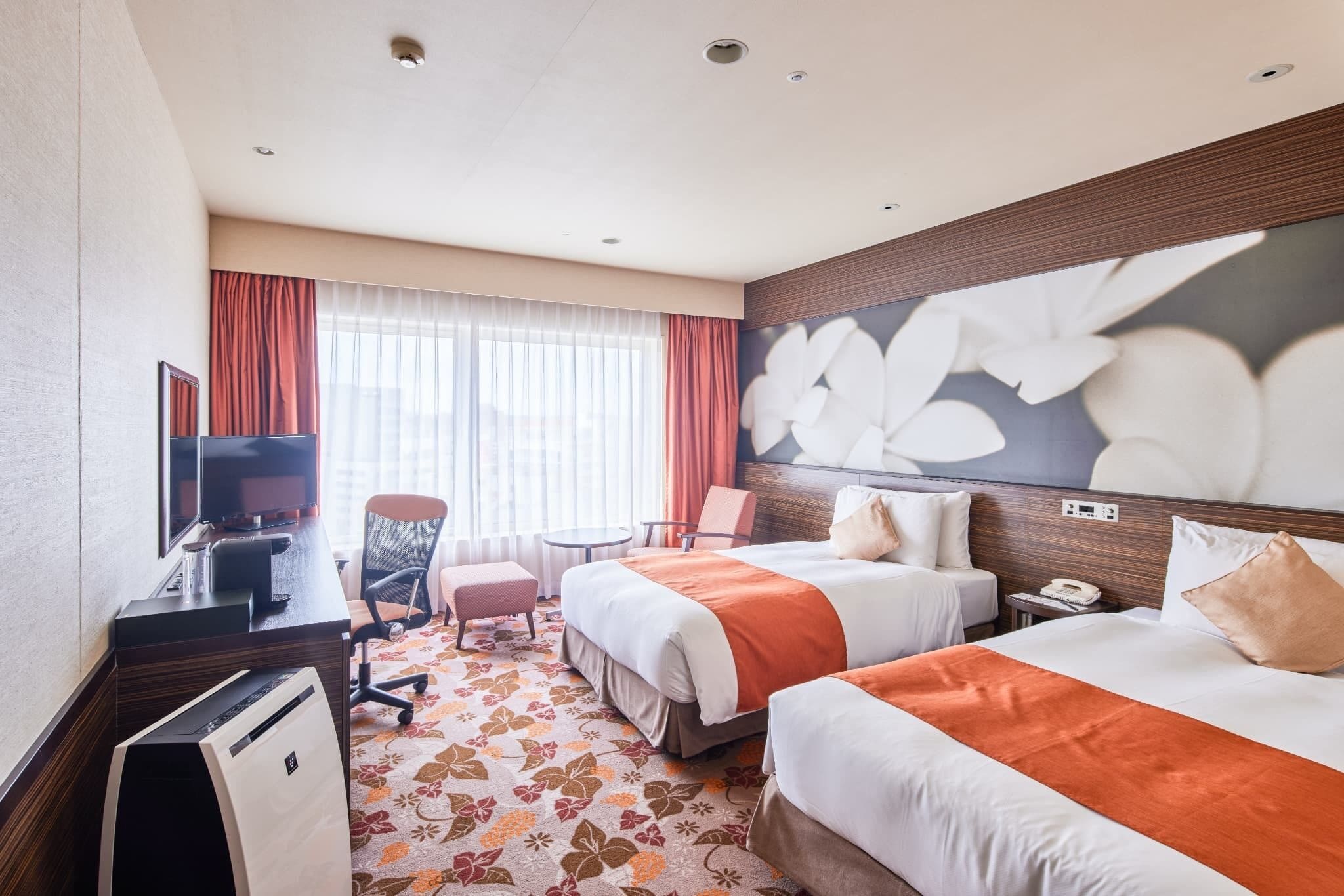 A hotel room with two beds, a desk with a computer, a chair, a TV, and a large floral wall mural.