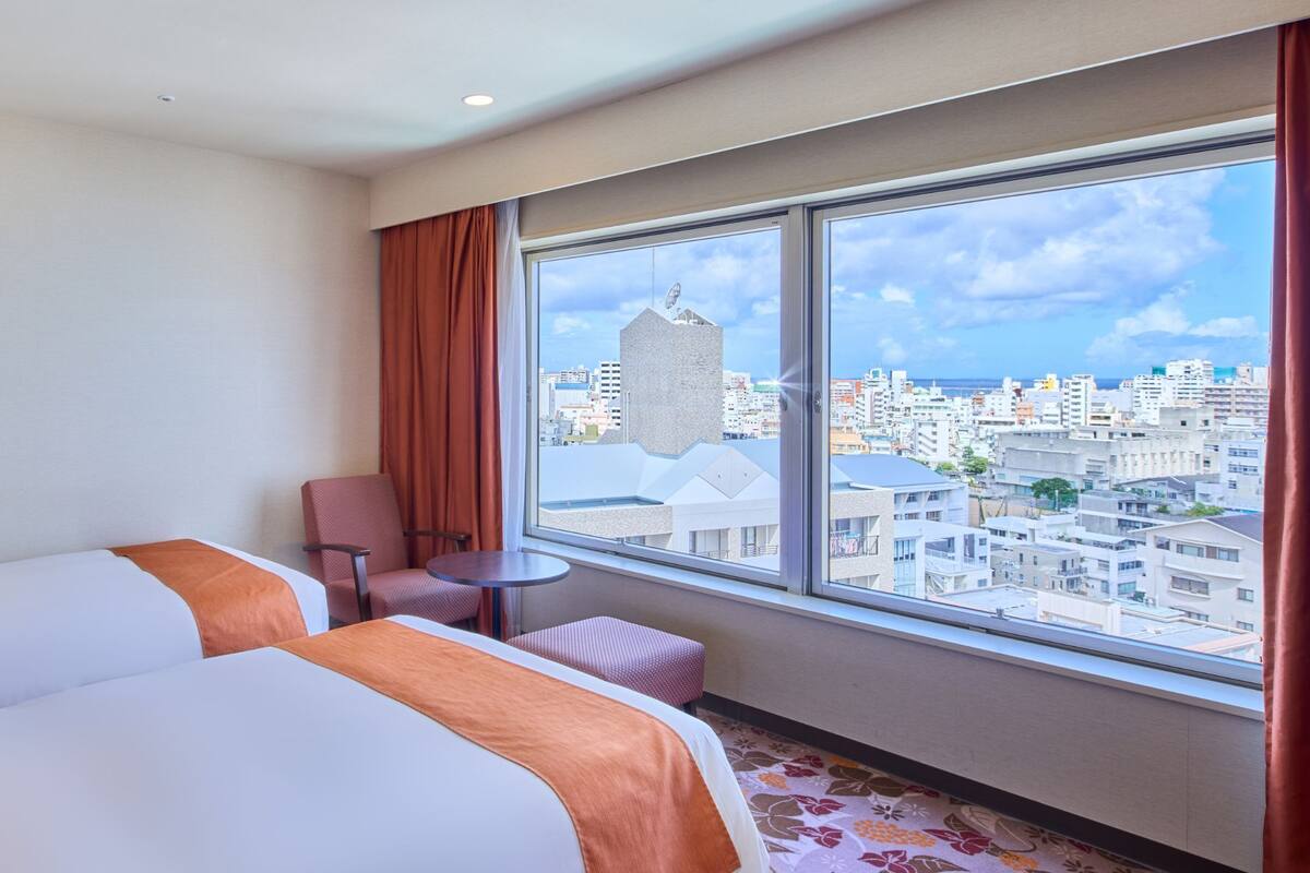 A hotel room with two beds, a small table, a chair, and a large window offering a city view.