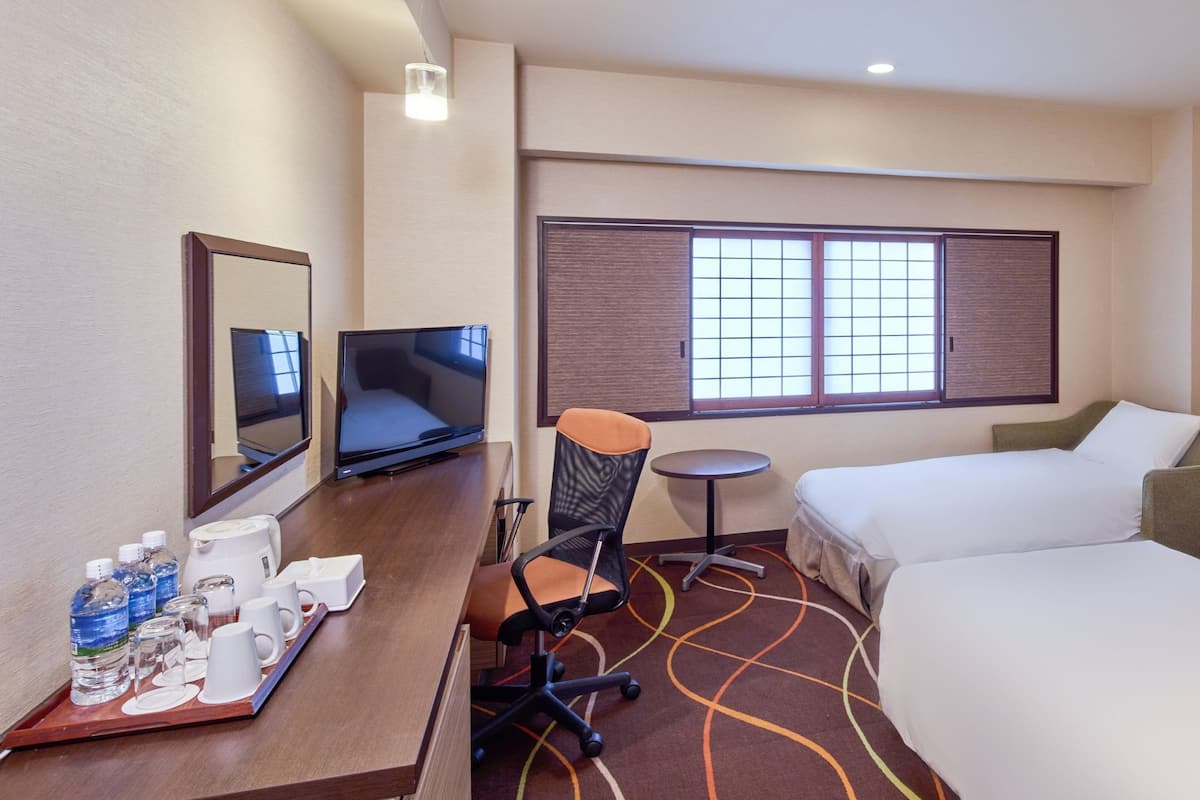 A hotel room with a bed, a desk with a chair, a television, a window with blinds, and a small table.