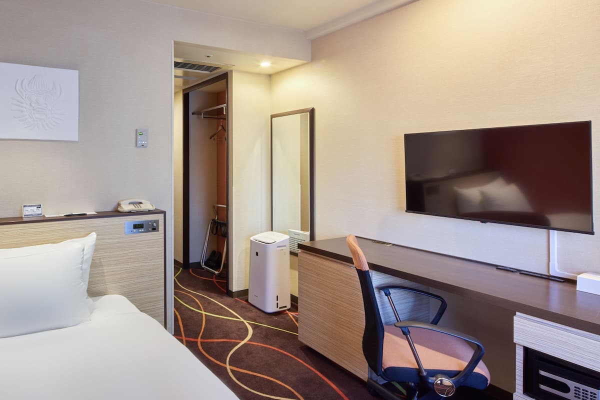 A hotel room with a bed, a desk with a chair, a TV, a mirror, and a door leading to a bathroom.