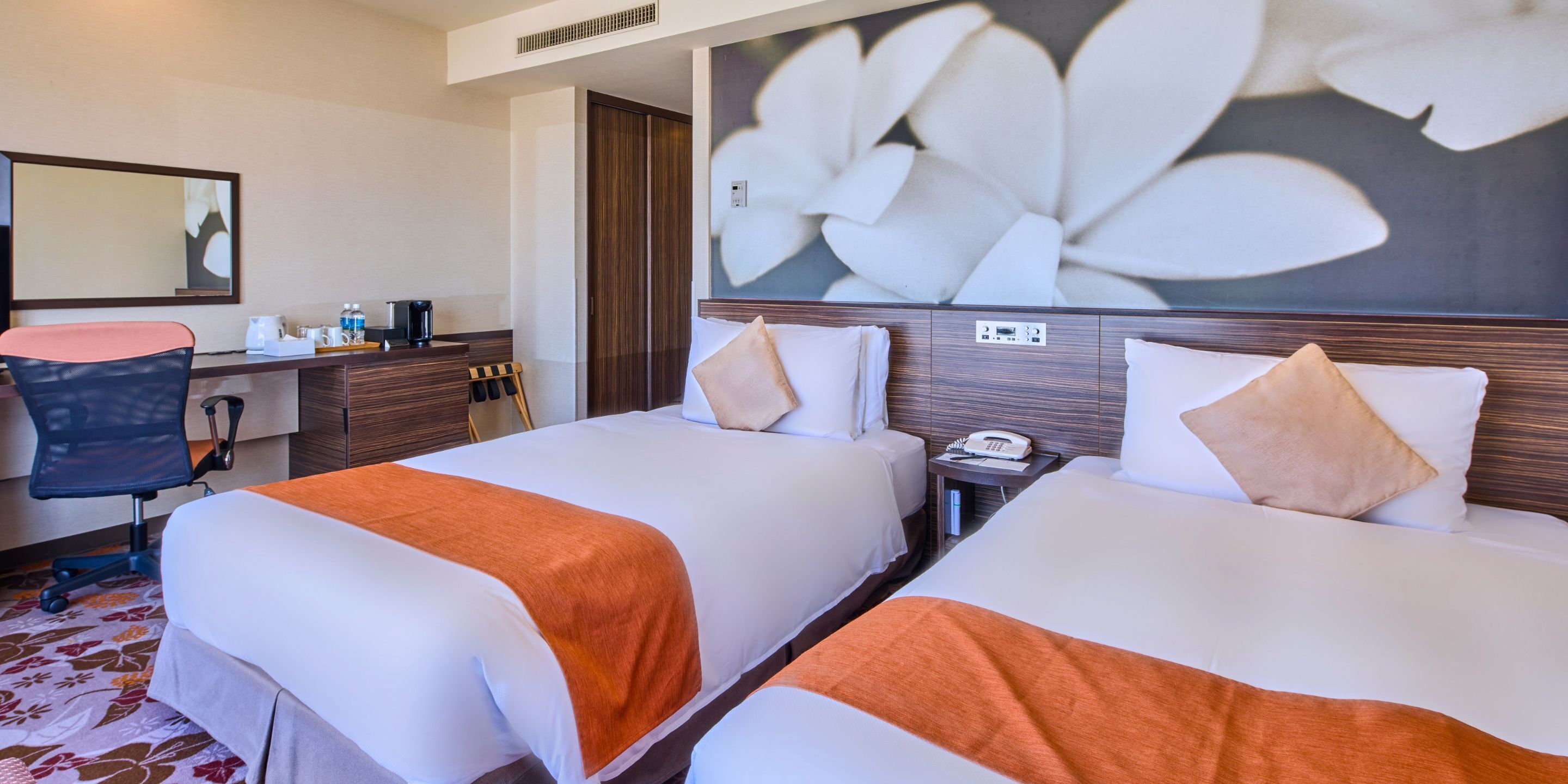A hotel room with two beds, a desk with a chair, a telephone, and a large floral artwork on the wall.