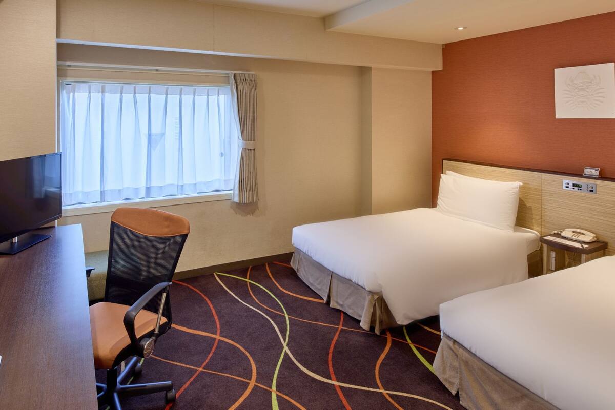 A hotel room with a desk, two beds, a TV, and a window with curtains.