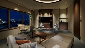 Een moderne woonkamer met een grote bank, twee fauteuils, een salontafel en een televisie. Een groot raam biedt uitzicht op de stad.