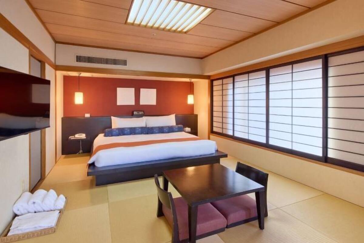 Une chambre d’hôtel de style japonais traditionnel, avec un sol recouvert de tatamis, un lit doté d’une tête de lit sombre, une table avec des coussins et des portes coulissantes de type shoji.