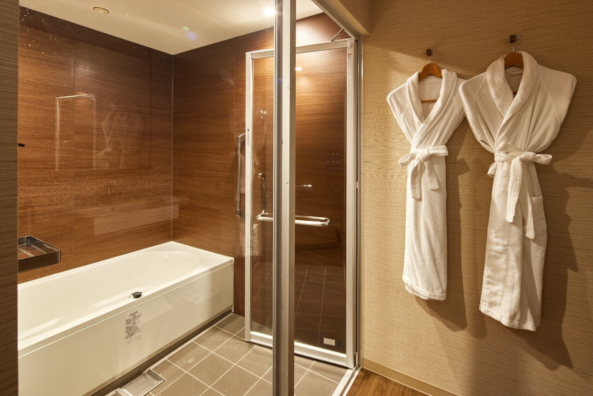 A modern bathroom with a glass-enclosed shower, a white bathtub, and two white robes hanging on the wall.