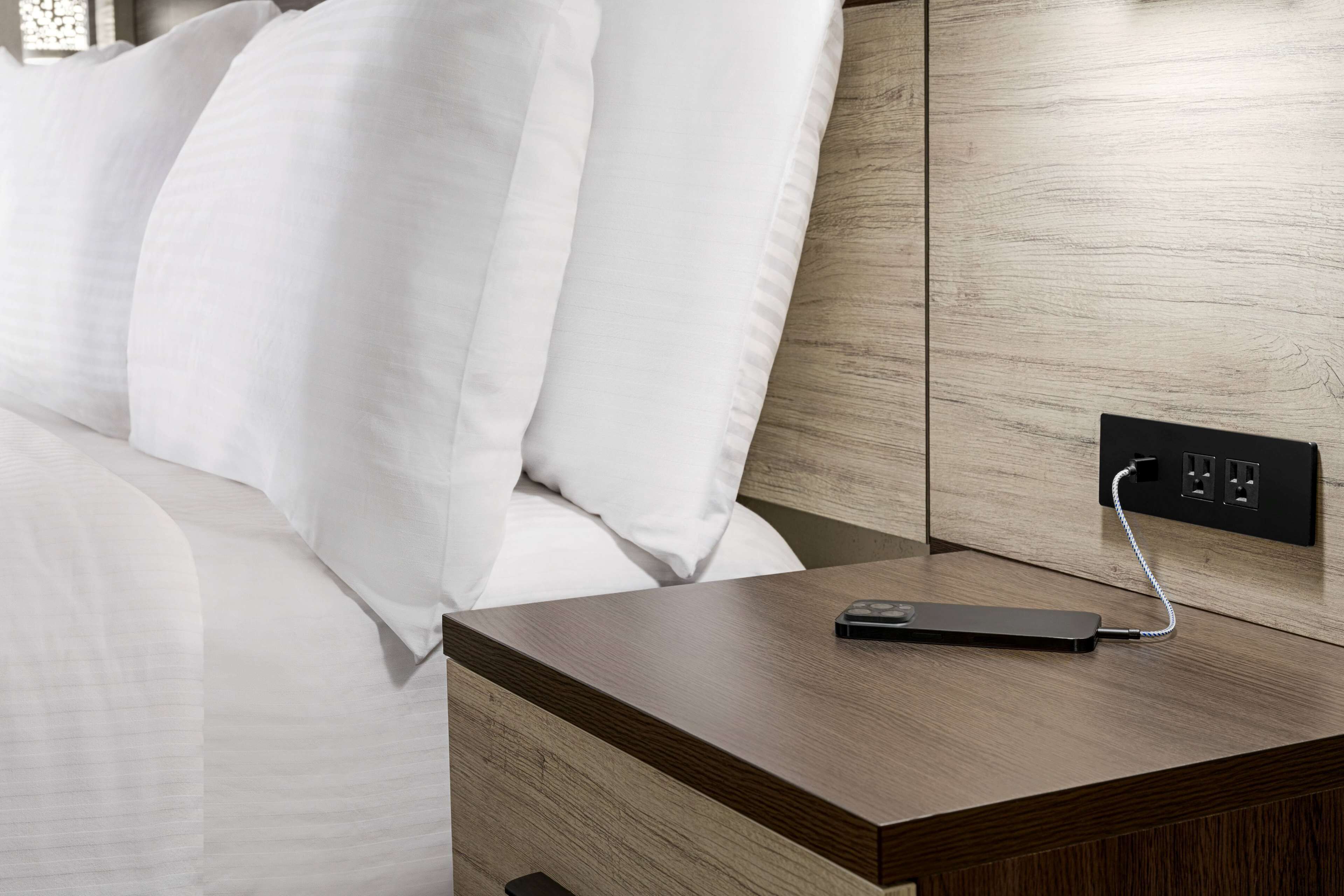 Una cama bien tendida con sábanas blancas, una mesita de noche de madera con un enchufe y un dispositivo de carga negro conectado.