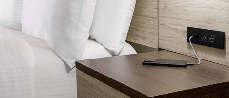 Una cama bien tendida con sábanas blancas, una mesita de noche de madera con un enchufe y un dispositivo de carga negro conectado.