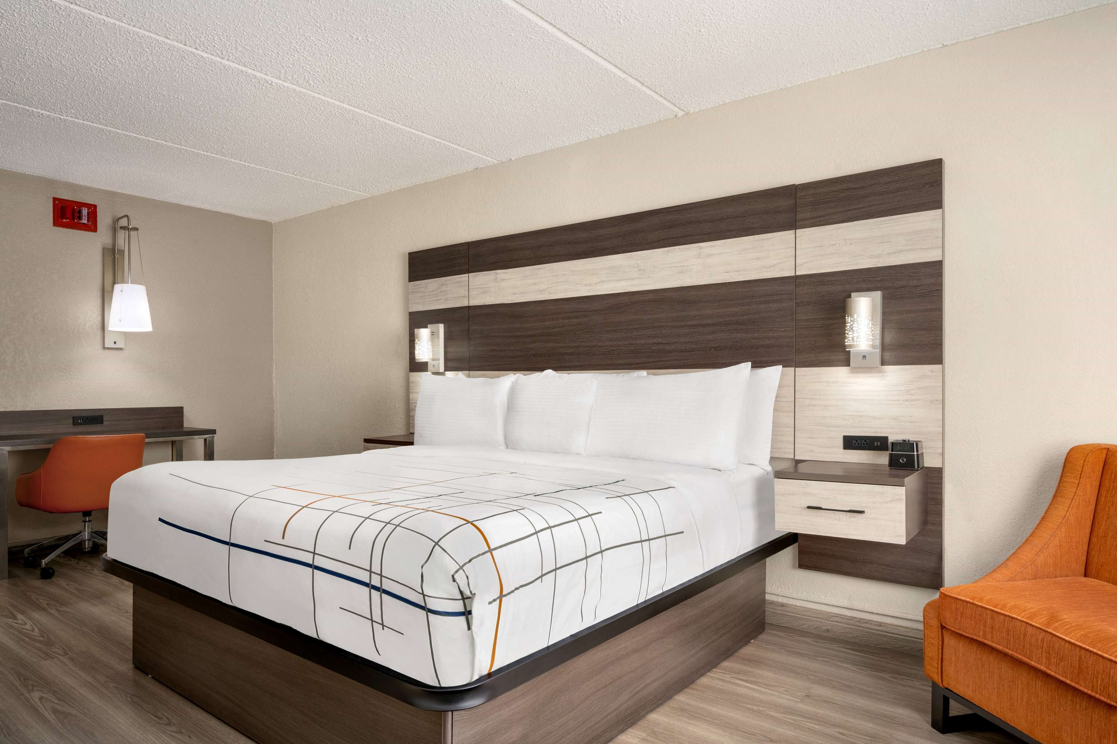 A modern hotel room with a large bed, a desk with a chair, and a nightstand with a lamp.