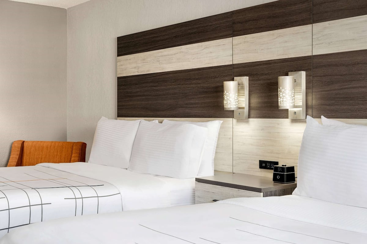A hotel room with two beds, a wooden headboard, a bedside table with a lamp, and a control panel.