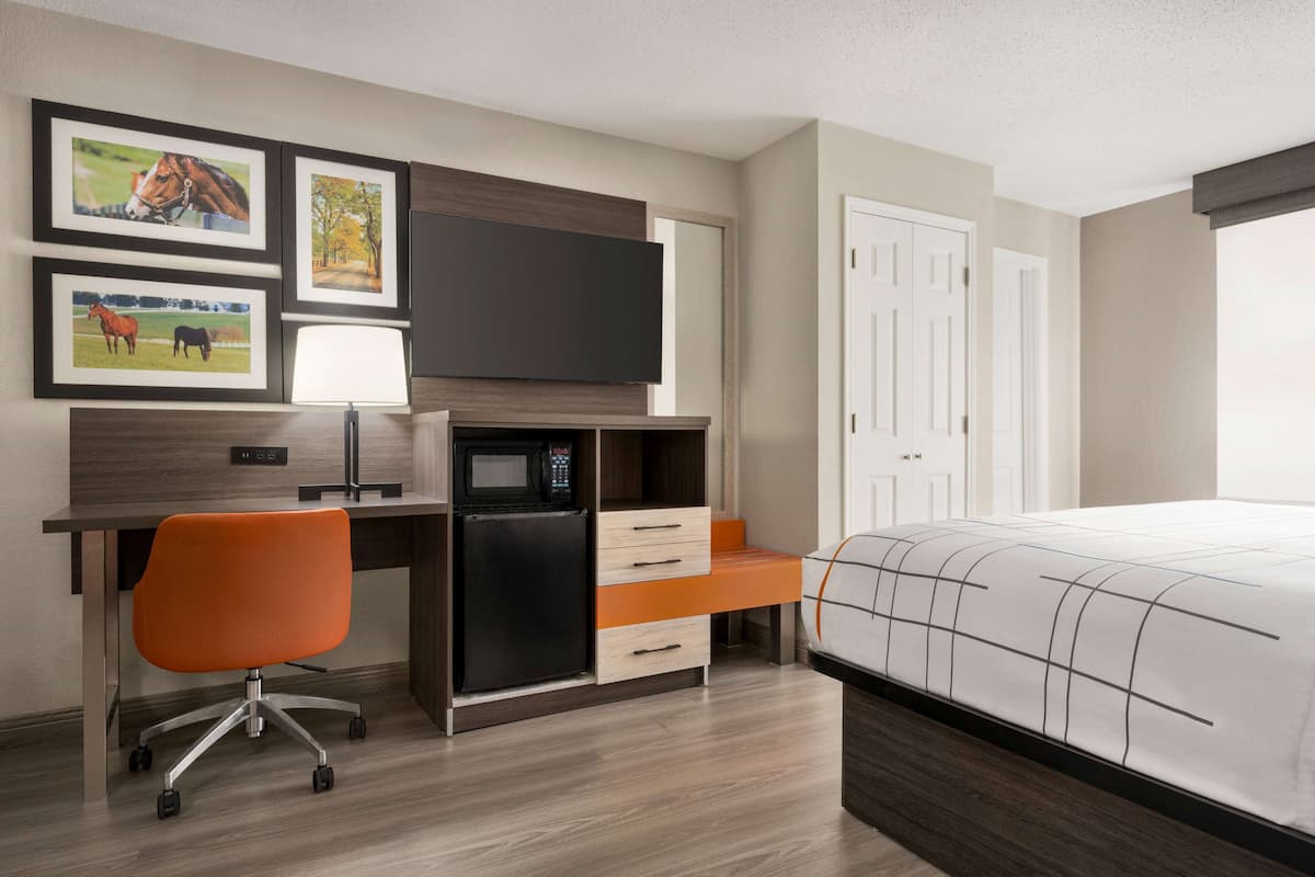 A hotel room with a bed, a desk with a computer, a microwave, and a TV. There are framed pictures on the wall and a door to the right.