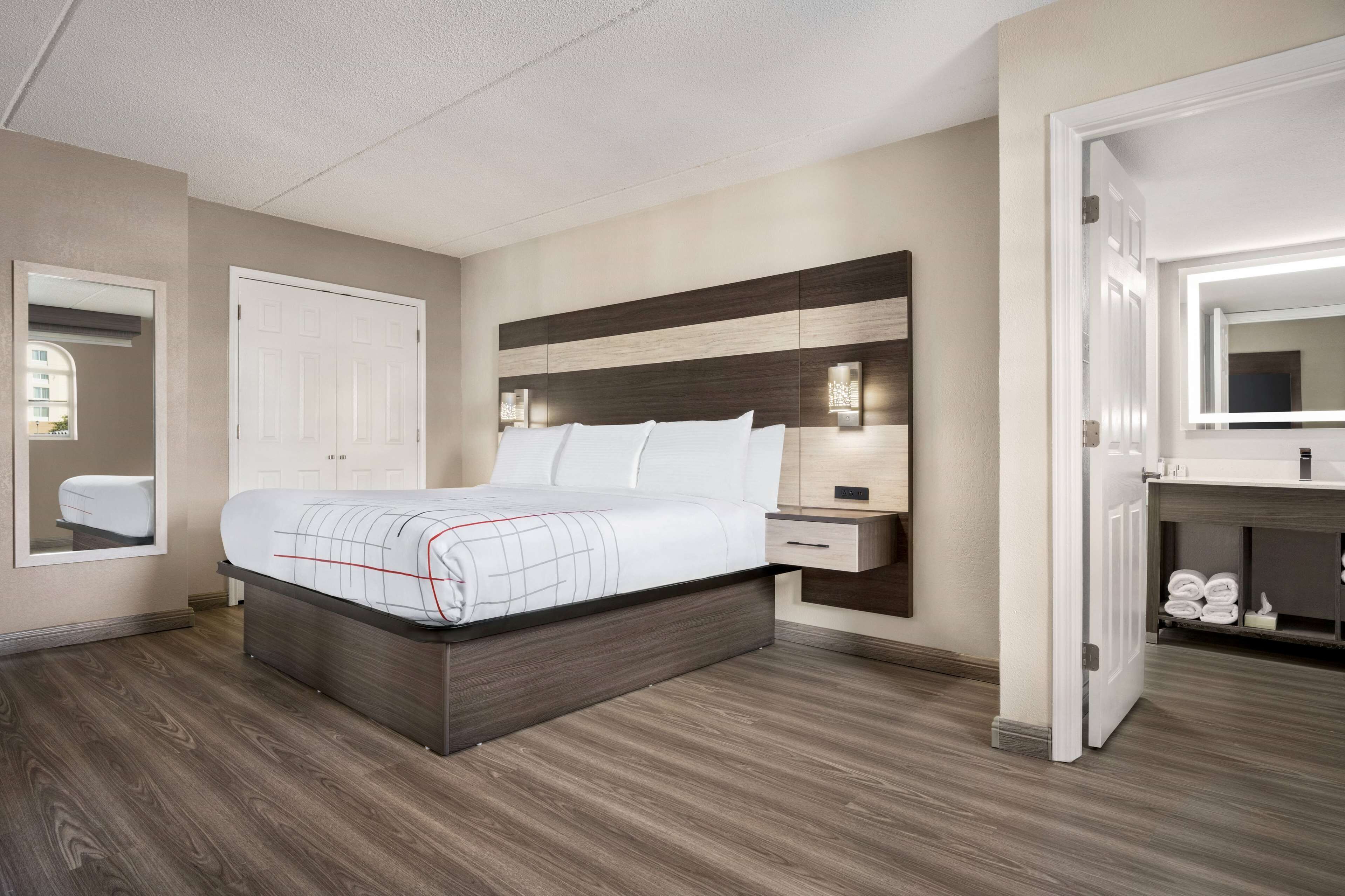 Une chambre d’hôtel avec un grand lit, une table de chevet, une commode, un miroir et une salle de bain équipée d’un lavabo et d’un porte-serviettes.
