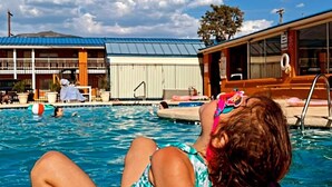 A pool area with a person sitting by the poolside, a building with a metal roof, and a clear blue sky. - The Classic Desert Aire Hotel (Alamogordo)