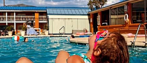 A pool area with a person sitting by the poolside, a building with a metal roof, and a clear blue sky.