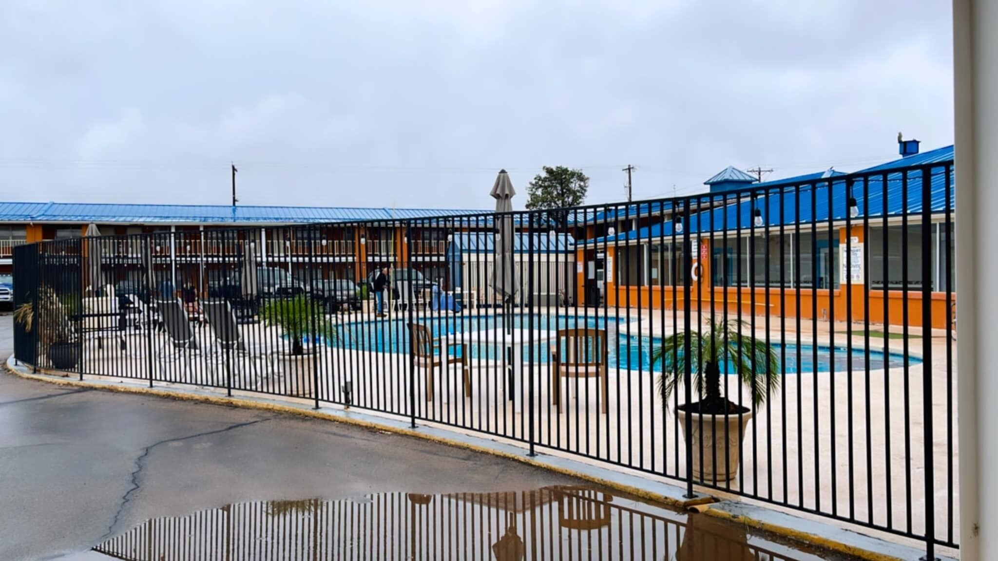 A fenced outdoor swimming pool area with a blue-roofed building in the background.