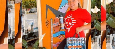 Una persona sentada en una silla con un bolso colorido, frente a una pared con un diseño geométrico.