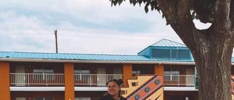 A person sitting on a wooden bench under a tree, with a building in the background.