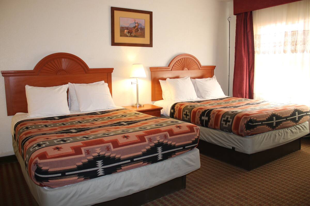 Une chambre d’hôtel avec deux lits, chacun recouvert d’une couvre-lit à motifs et agrémenté de coussins blancs. Un tableau encadré est accroché au mur au-dessus des lits, et une lampe sur pied se trouve entre les deux.