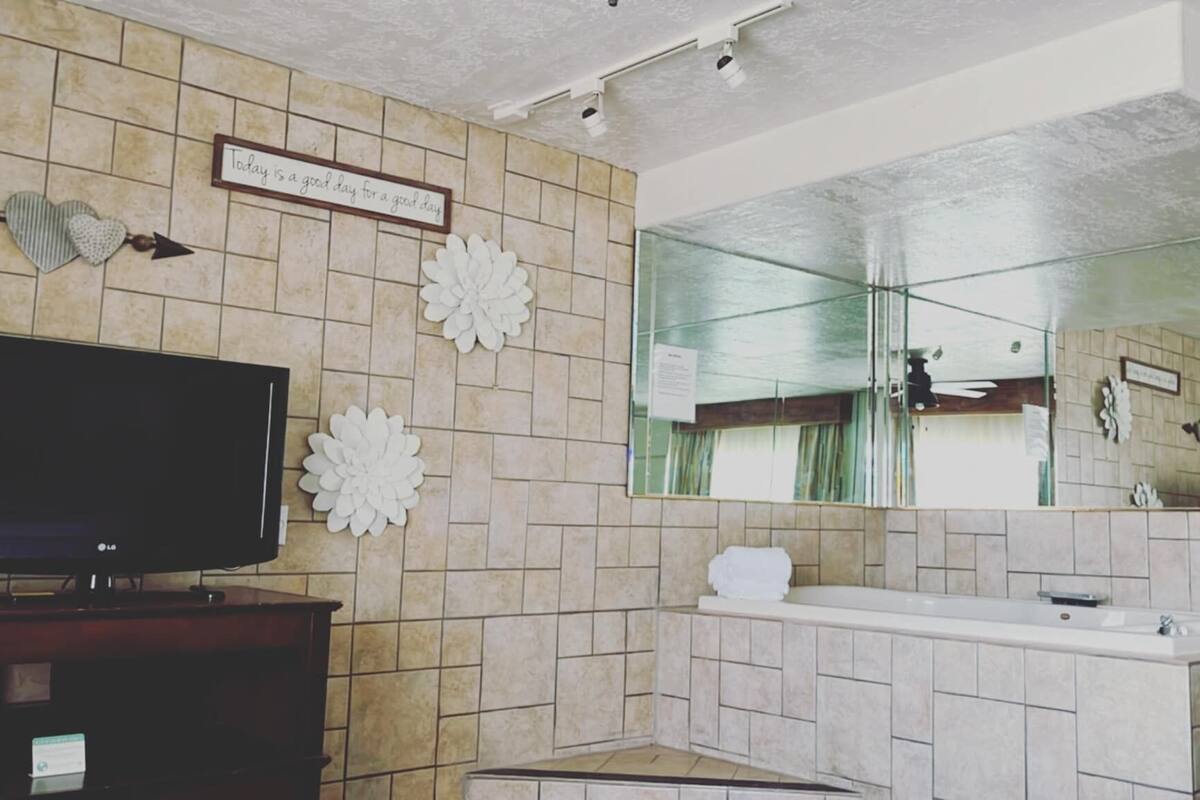 Une salle de bain avec un mur carrelé, un grand miroir, un lavabo et un téléviseur posé sur un meuble.