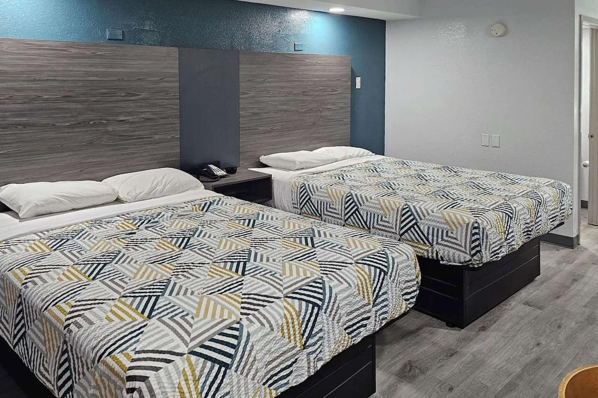 A hotel room with two beds, each with a patterned bedspread and white pillows. The room has a modern design with dark wood headboards and a grey accent wall.