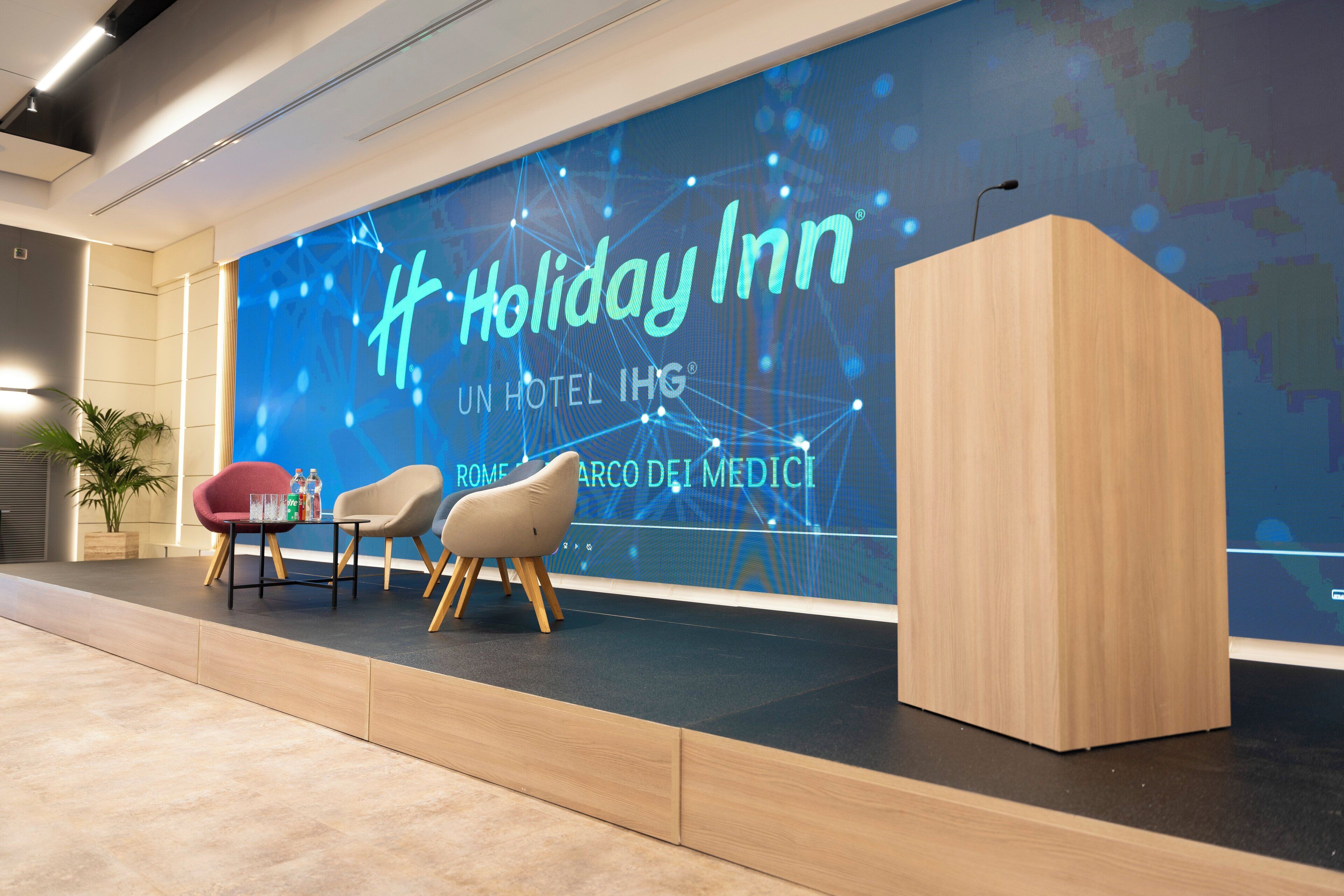 A modern hotel lobby with a large digital display reading "Holiday Inn" and "Rome - Marco dei Medici". There are two chairs and a table with water bottles.