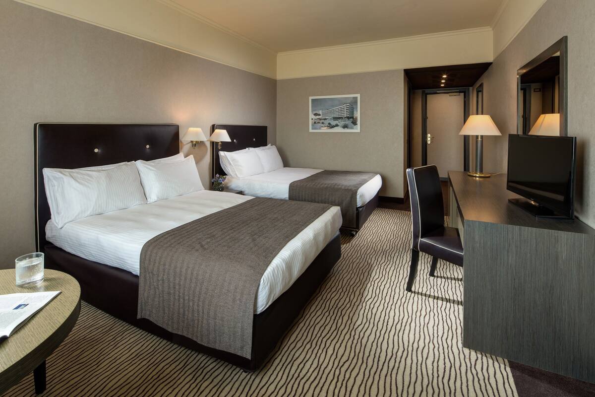 A hotel room with two beds, a desk, a chair, a TV, and a painting on the wall.