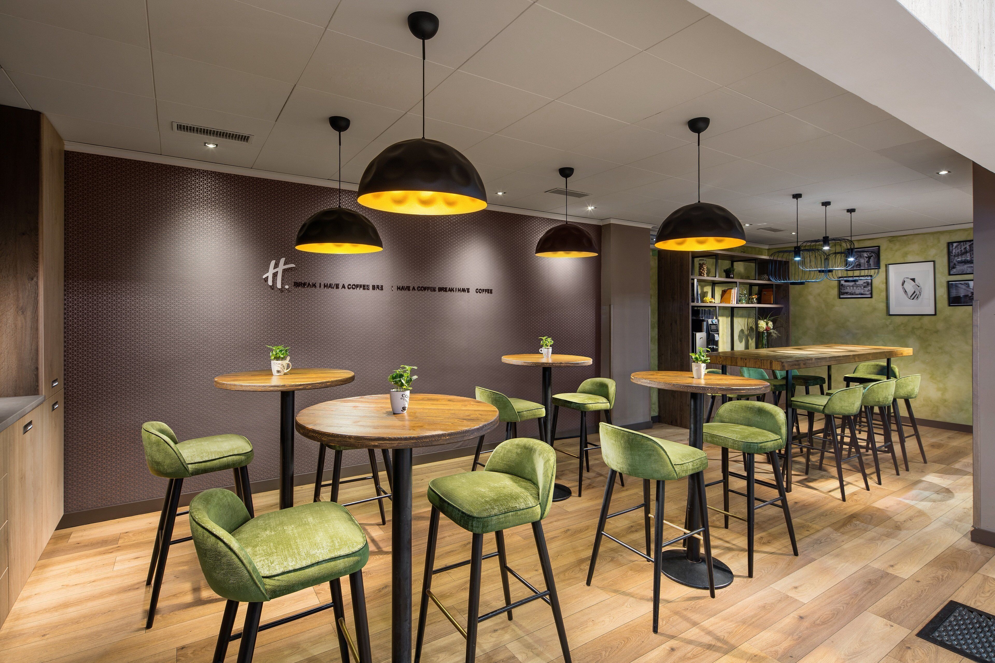 L’intérieur d’un café moderne, avec des tables rondes, des chaises recouvertes d’un tissu vert et des luminaires suspendus.