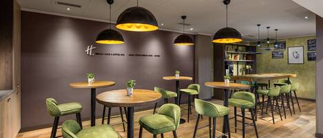 L’intérieur d’un café moderne, avec des tables rondes, des chaises recouvertes d’un tissu vert et des luminaires suspendus.
