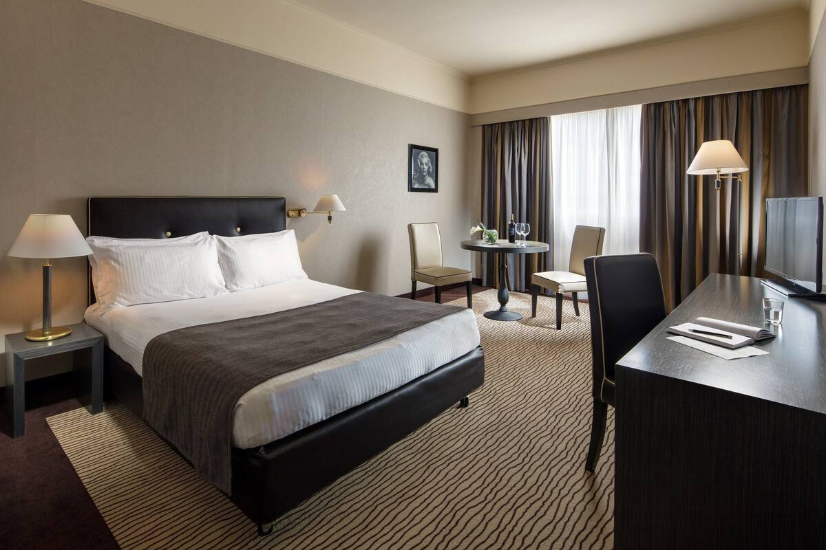 A hotel room with a large bed, a desk, a chair, a small table with a lamp, and a television.