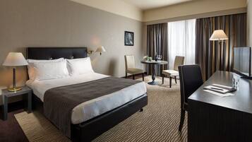 A hotel room with a large bed, a desk, a chair, a small table with a lamp, and a television.