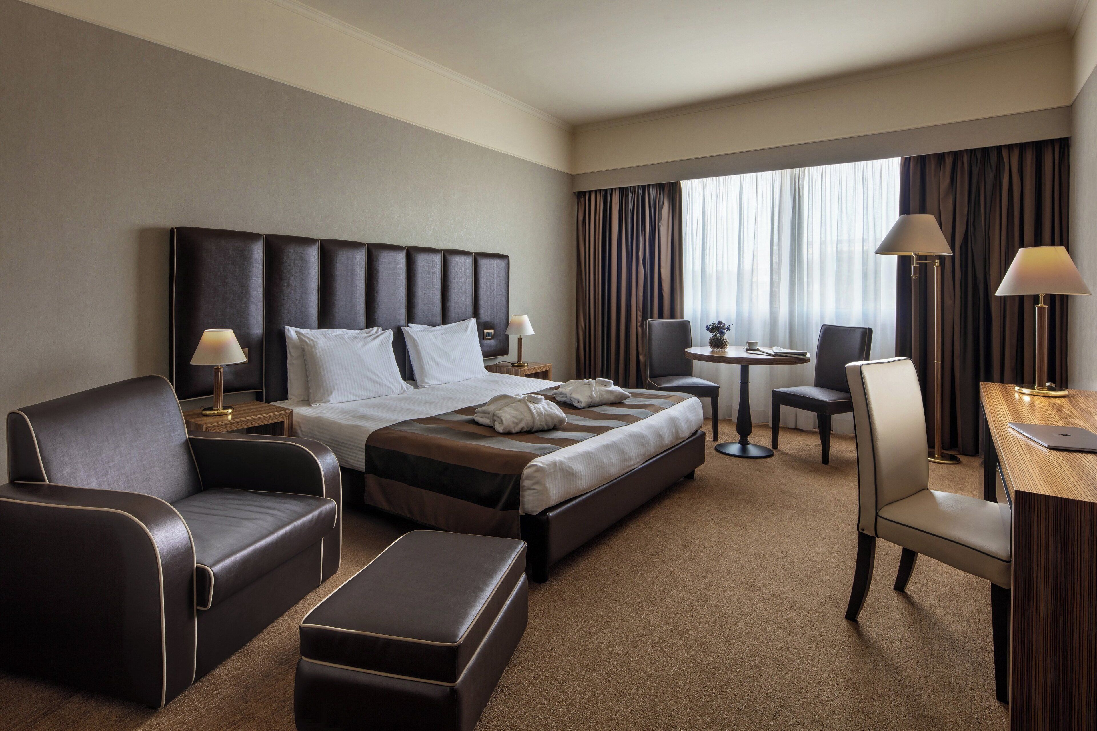 Quarto de hotel com uma cama grande, duas poltronas, uma escrivaninha com cadeira, uma mesinha e uma janela com cortinas.