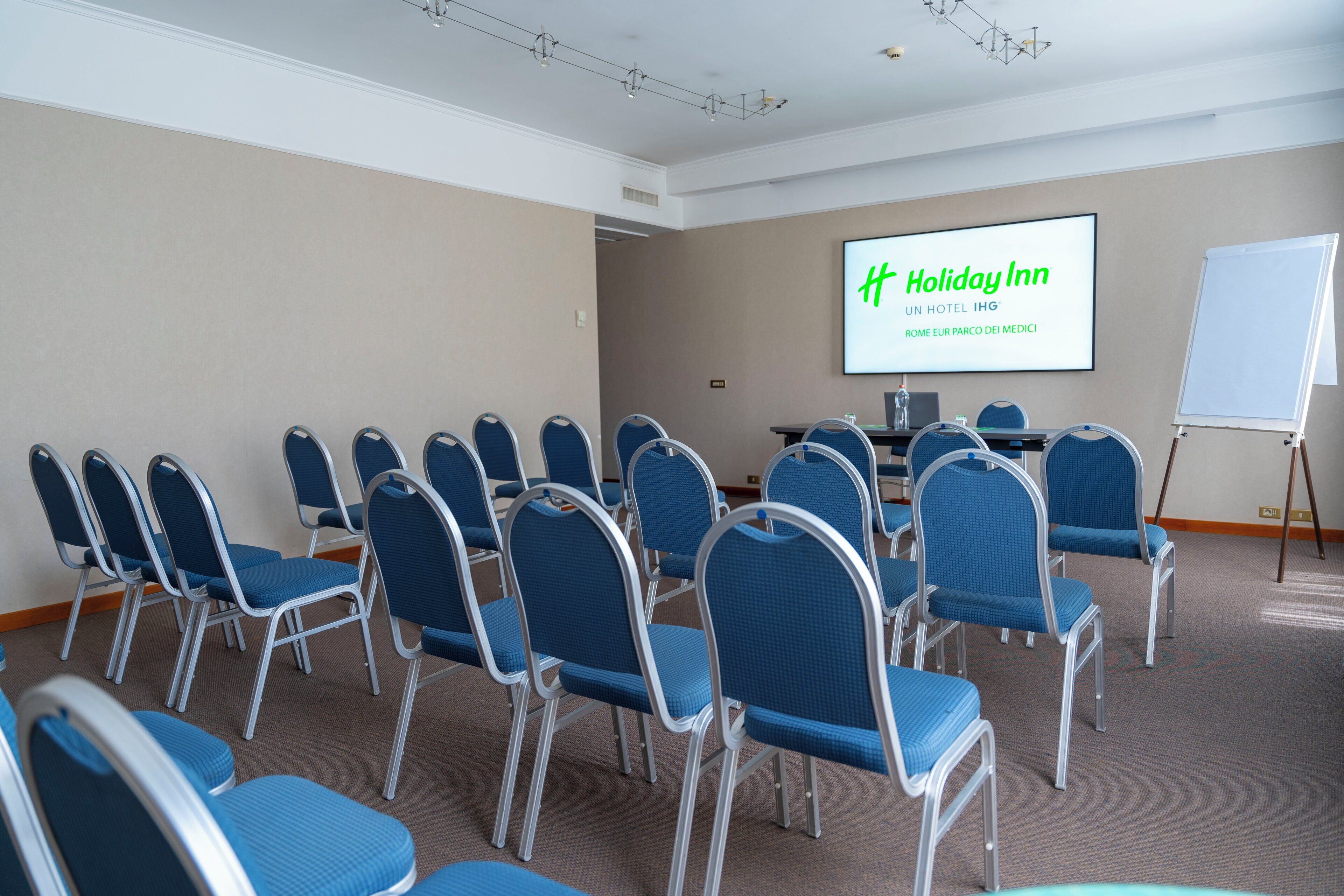 Una sala conferenze con file di sedie blu e un grande schermo che mostra il marchio Holiday Inn.