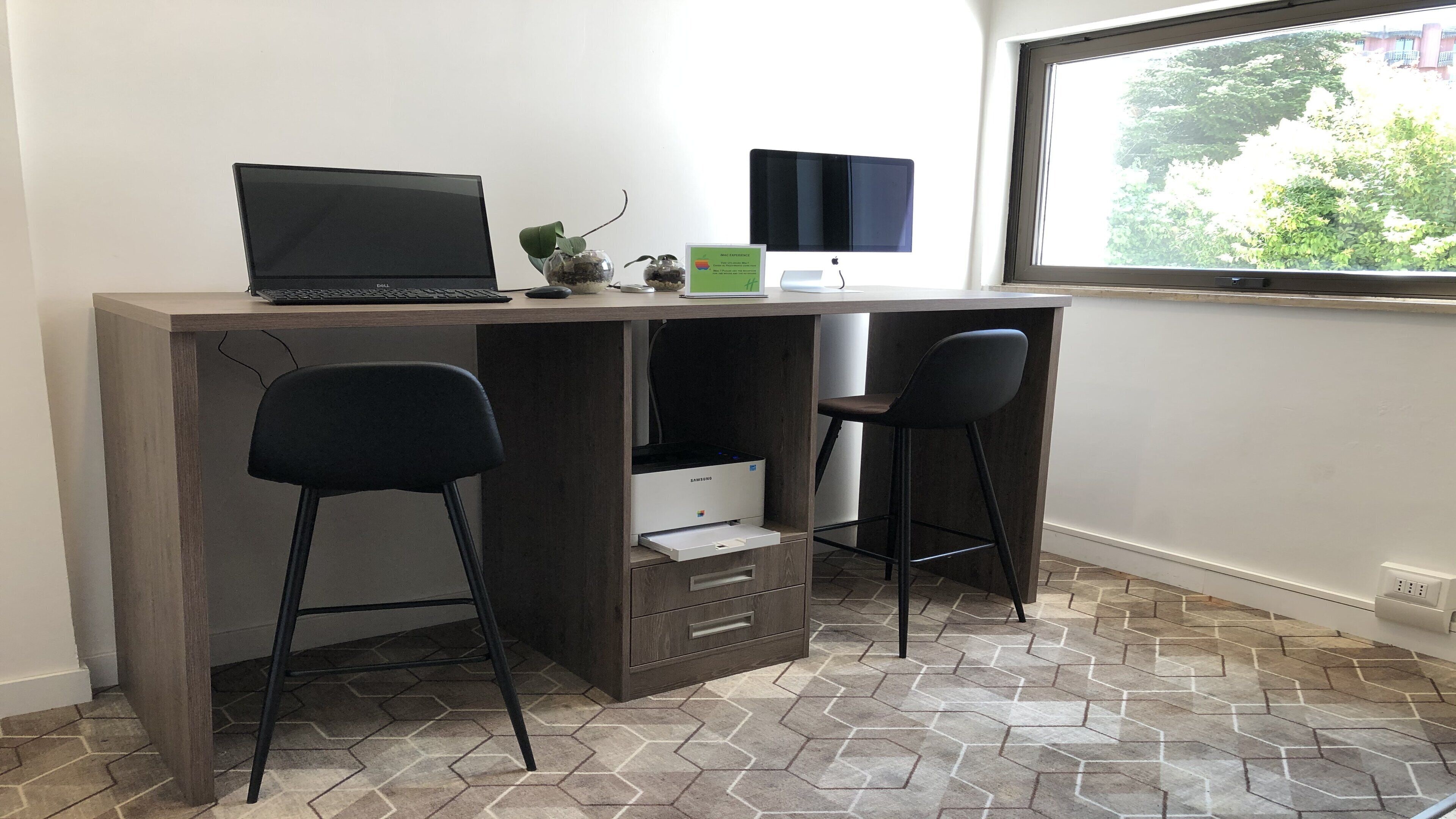 A modern workspace with a desk, two black chairs, a laptop, a printer, and a window with a view of greenery.