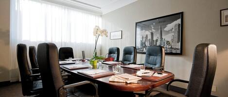 A conference room with a long table, multiple chairs, a potted plant, and a cityscape painting on the wall.