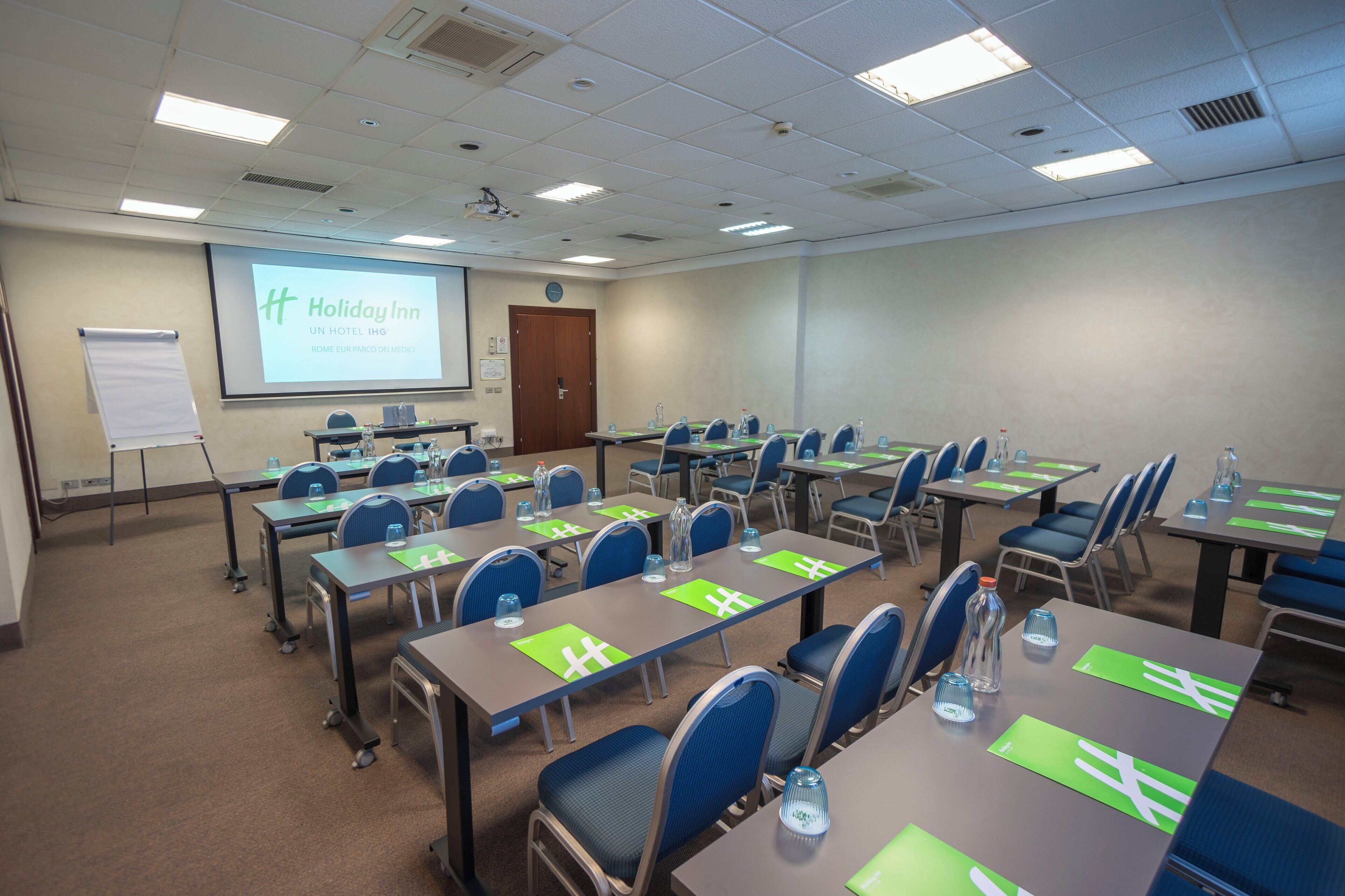 Ein Konferenzraum mit mehreren Tischen und Stühlen, einer Projektionsleinwand mit dem Schriftzug 'Holiday Inn' und einer Whiteboard-Tafel.