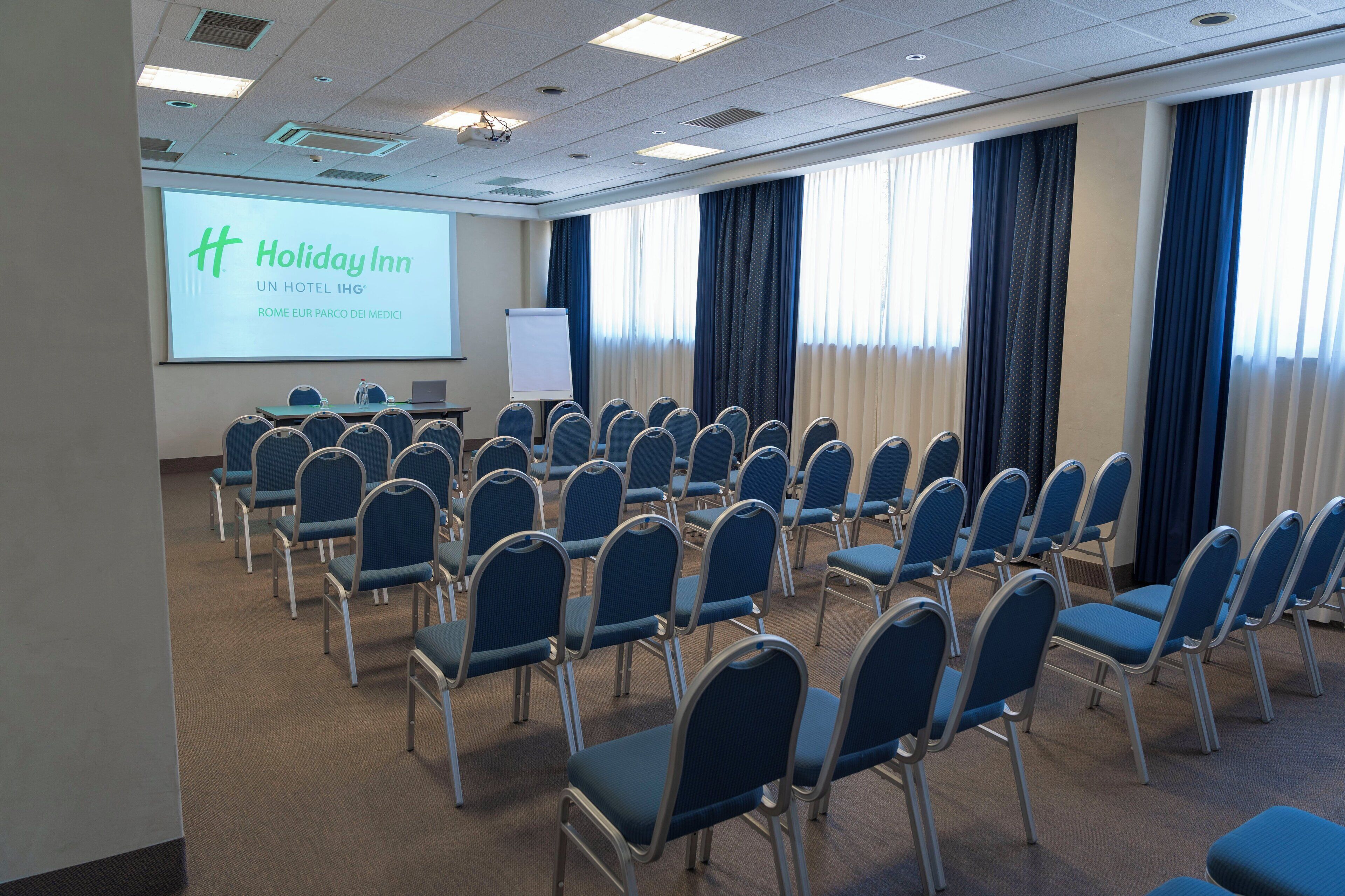 A conference room with rows of blue chairs, a projection screen displaying 'Holiday Inn', and a whiteboard.